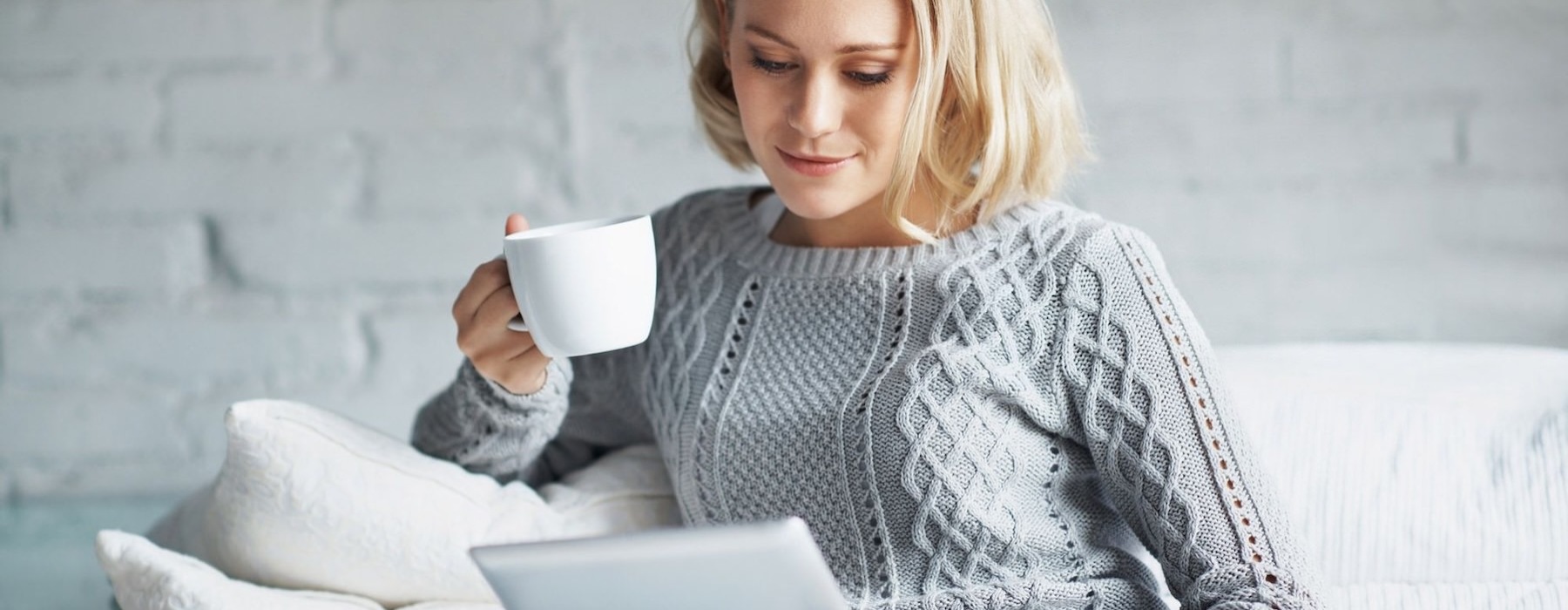 a woman sits on a couch with a cup of coffee and looks at her tablet