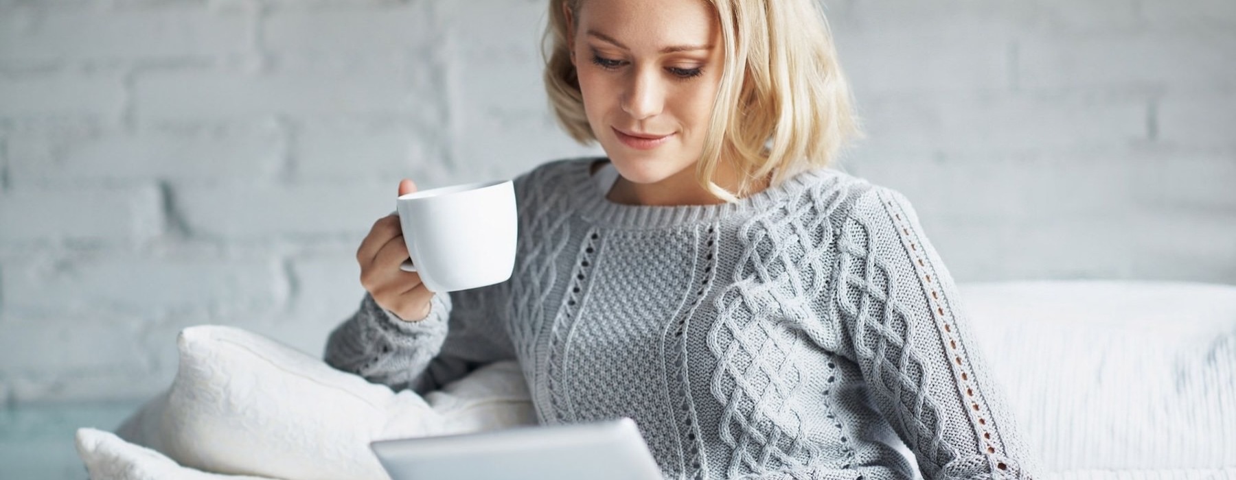 a woman sits on a couch with a cup of coffee and looks at her tablet